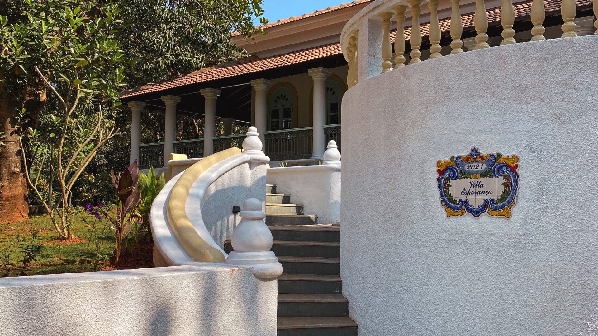 A picturesque view of the Villa Esperança entrance, featuring a sweeping white staircase and traditional Indo-Portuguese architecture nestled amidst lush greenery.