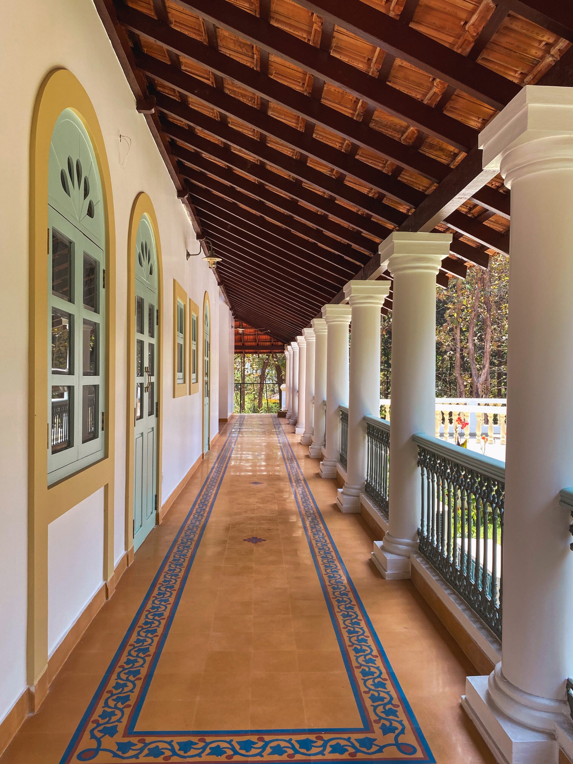 An expansive Indo-Portuguese style veranda featuring a rhythmic colonnade, exposed wooden roof trusses, and heritage floor tiles with intricate blue borders.