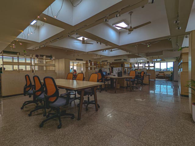 Open-Plan Synergy: The expansive workspace is designed to foster collaboration, featuring modular desks and vibrant orange seating under a high, skylit ceiling.